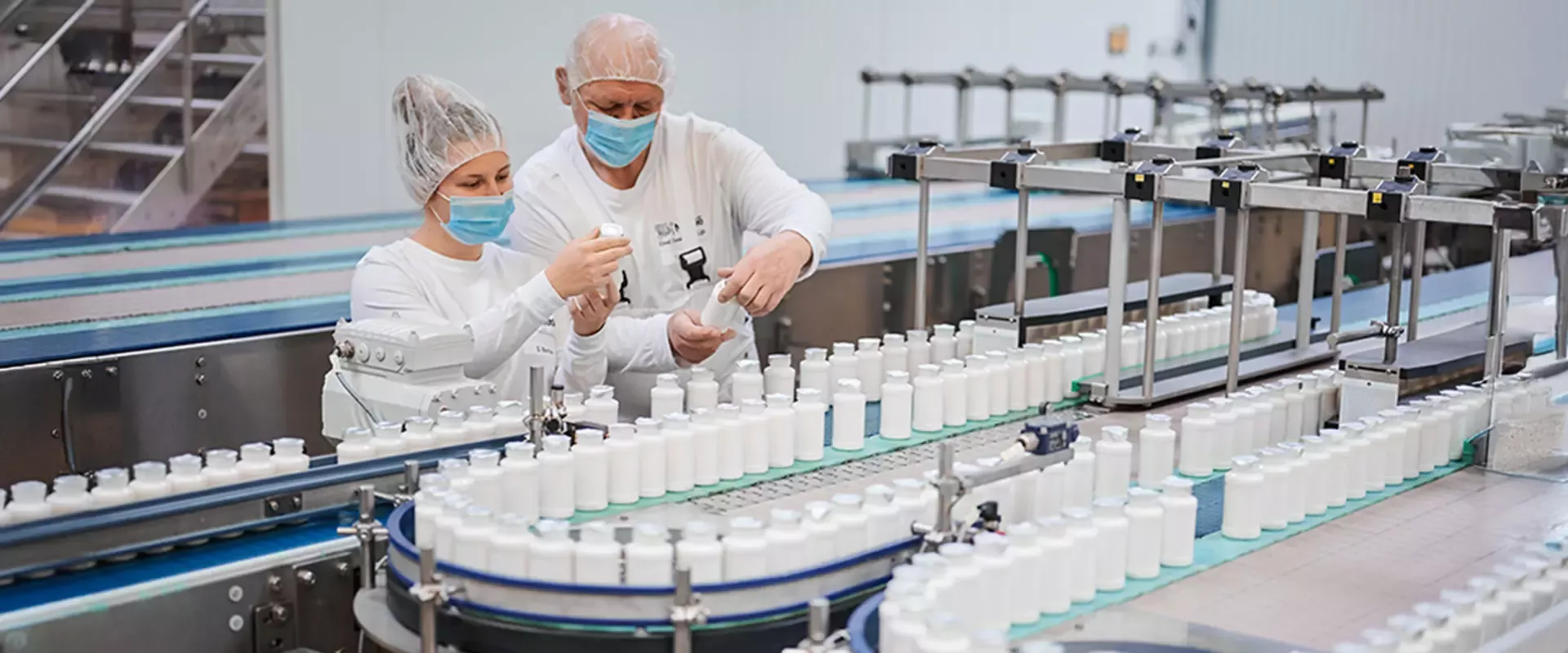 Zwei Arbeiter, die Haarnetze und Masken tragen, prüfen Flaschen auf einem Förderband in einer sauberen, modernen Fabrik, was Präzision und Qualitätsbewusstsein vermittelt.