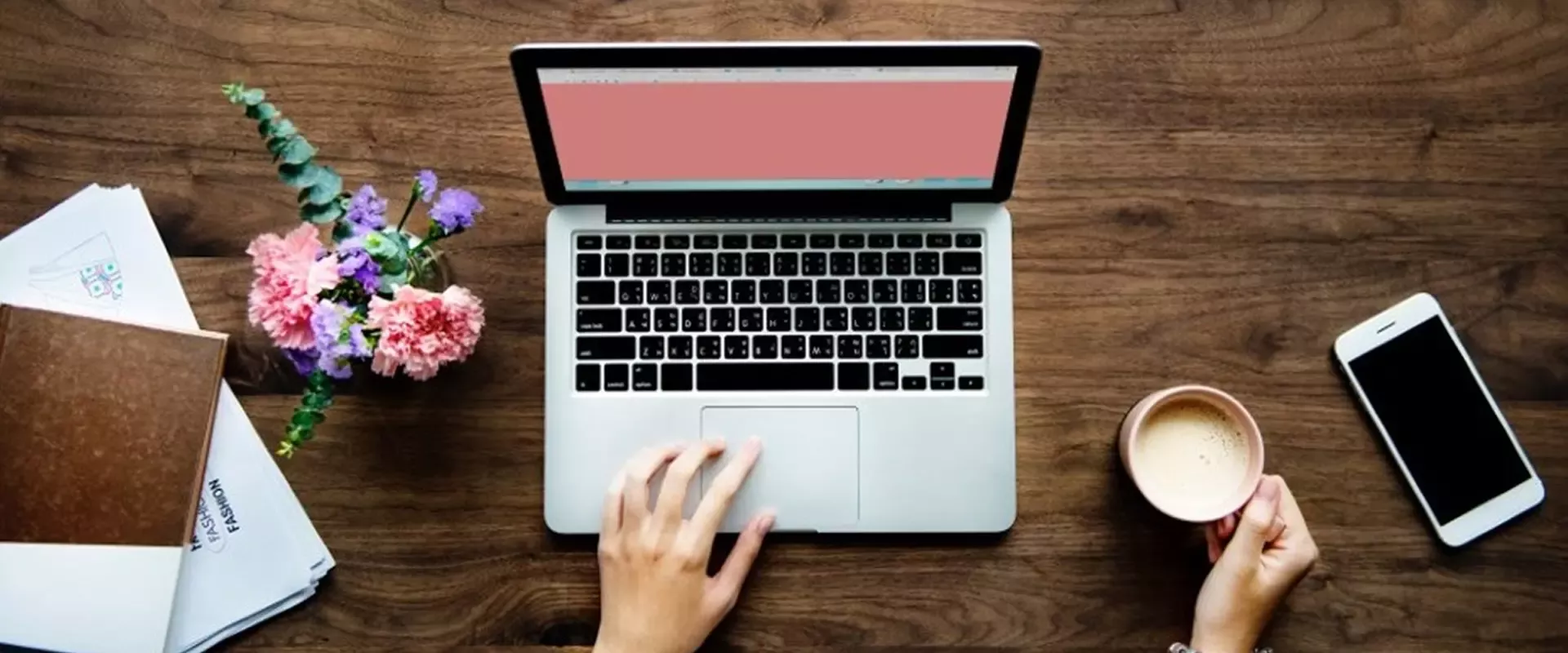 Eine Person arbeitet an einem Laptop mit einem leeren rosa Bildschirm, die eine Hand auf der Tastatur und in der anderen eine Kaffeetasse. Daneben liegen ein Telefon, ein Notizblock und bunte Blumen.