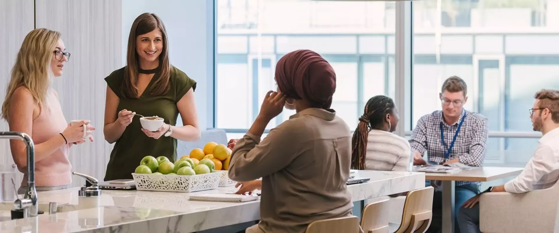 Eine Gruppe von Menschen versammelt sich in einer hellen, modernen Büroküche. Drei Frauen unterhalten sich an der Kücheninsel, auf der Obst liegt, während vier Personen am Tisch sitzen und diskutieren.