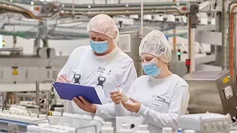 Zwei Arbeiter in Schutzkleidung und mit Gesichtsmasken prüfen Flaschen in einer Fabrikhalle.